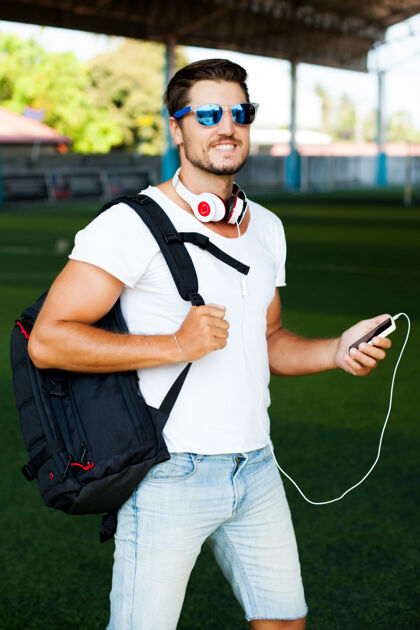 肌肉男年轻时尚的男子在足球场上摆姿势 听音乐头戴耳机 手拿球员 脸戴墨镜背包玻璃肖像