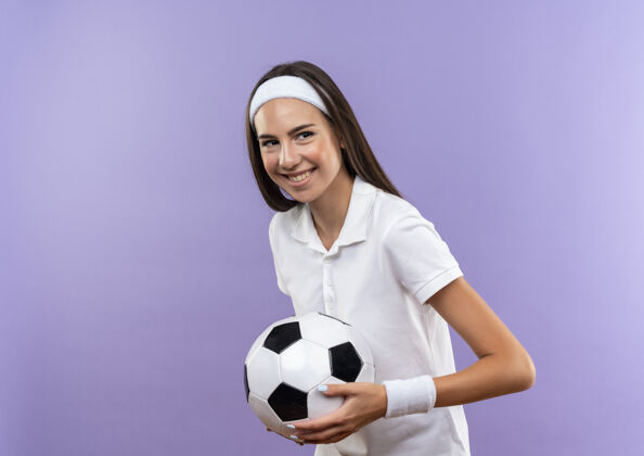 运动微笑着漂亮的运动女孩戴着头带和手环拿着足球孤立在紫色的空间女孩微笑漂亮