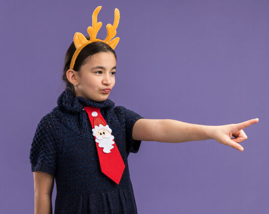 索引穿着针织连衣裙的小女孩戴着红领带 边上有滑稽的边 头上有鹿角 她用食指指着什么东西圣诞旁边有趣