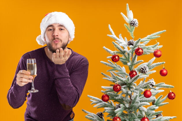吹着穿着紫色毛衣 戴着圣诞帽的年轻人在橙色背景下的圣诞树旁举着一杯香槟杯子男人圣诞树