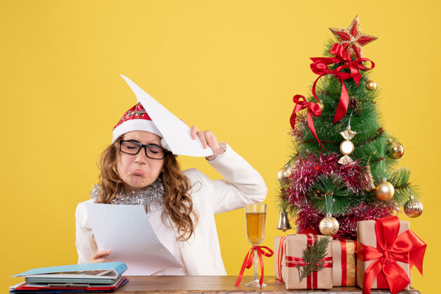 圣诞节前视图女医生坐在圣诞树上 拿着黄色背景上的文件快乐背景礼物