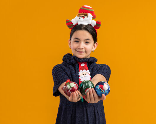 快乐快乐的小女孩穿着针织连衣裙 头上系着一条红色的领带 头上戴着一个滑稽的边 手里拿着圣诞球 脸上带着微笑 看上去快乐而积极搞笑举行头