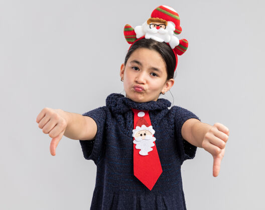 头小女孩穿着针织连衣裙 头上系着一条红色领带 上面戴着一个有趣的圣诞戒指 看起来很不高兴 竖起大拇指针织新搞笑