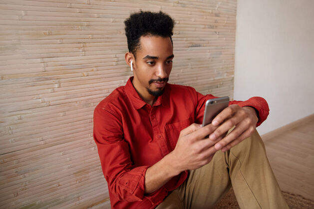 专注专注的年轻黑发男性 皮肤黝黑 双手举着智能手机 坐在米色内饰的地板上 专心地看着屏幕心情皮肤智能手机