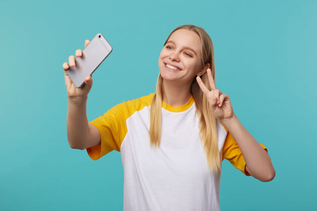 高兴快乐的年轻可爱的金发长发女人在手机上展示胜利的姿态 穿着休闲装摆出蓝色的姿势长肖像黄色