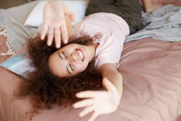 美国年轻开朗的非洲裔美国妇女在家里享受阳光明媚的日子 在家里度过空闲的一天 躺在床上 闭着眼睛 举起手 面带微笑学生快乐脸