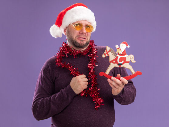 中年皱眉的中年男子戴着圣诞老人帽 脖子上戴着金箔花环 戴着眼镜 把圣诞老人抱在紫色墙上的摇马雕像上圣诞老人摇摆围着