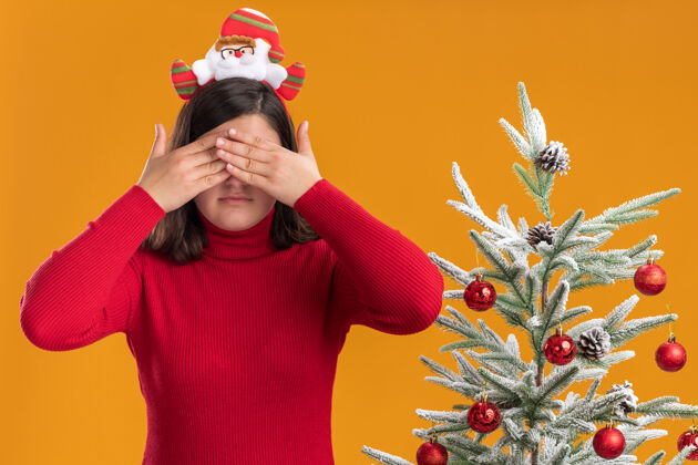 眼睛穿着圣诞毛衣的年轻女孩戴着有趣的头带 手放在橙色背景下的圣诞树旁遮住眼睛站着毛衣年轻