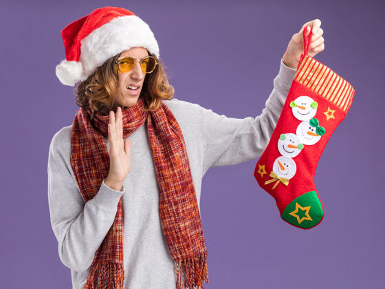 表情戴着圣诞老人帽 戴着黄色眼镜 脖子上围着暖和的围巾 手里拿着圣诞长袜 站在紫色背景下 带着厌恶的表情看着它温暖围着快乐