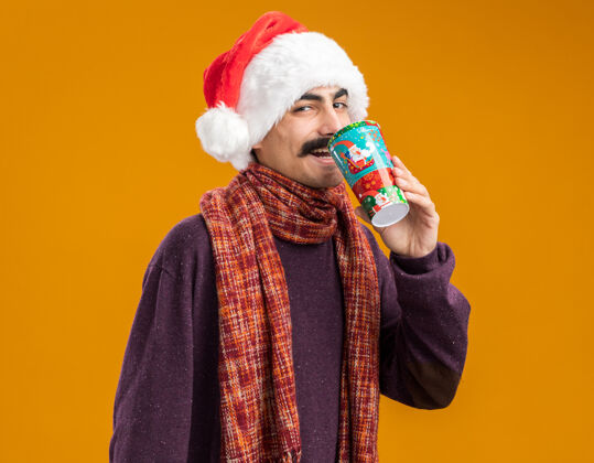 圣诞快乐留着胡子的男人戴着圣诞老人帽 脖子上围着暖和的围巾 喝着五颜六色纸杯里的果汁 站在橙色的背景下 快乐而愉快围着围巾脖子