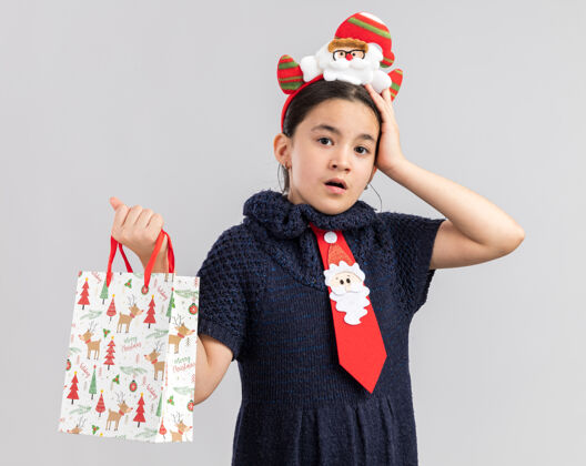 拿着小女孩穿着针织连衣裙 头上系着一条红色领带 头上戴着一个有趣的圣诞边 手里拿着一个装有圣诞礼物的纸袋 看上去很困惑 手放在头上新年纸手