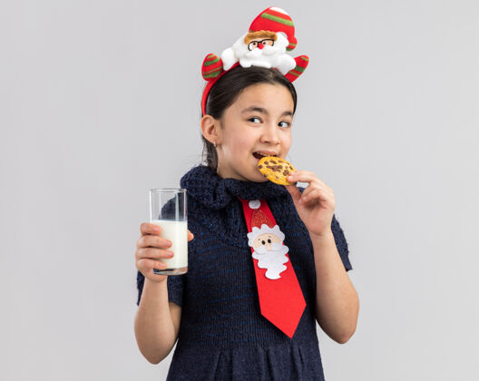 新年快乐的小女孩穿着针织连衣裙 头上系着一条红色领带 头上戴着一个有趣的圣诞戒指 手里拿着一杯牛奶 吃着饼干领带拿着快乐