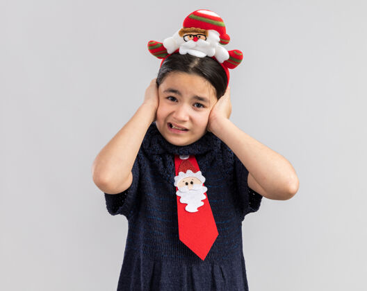 领带小女孩穿着针织连衣裙 头上系着一条红色领带 戴着滑稽的圣诞戒指 手捂着耳朵 表情烦躁小衣服边