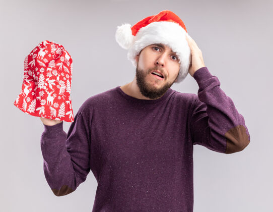 错误一个穿着紫色毛衣 戴着圣诞帽的年轻人 手里拿着一个红色的礼物包 看上去很困惑 因为他站在白墙上的时候犯了个错误圣诞节头男人