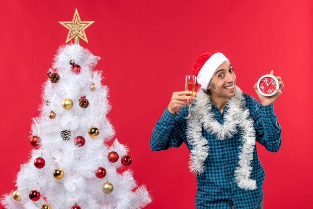 年轻人快乐的年轻人戴着圣诞老人的帽子 拿着一杯葡萄酒和一个红色圣诞树旁的钟圣诞圣诞老人时钟