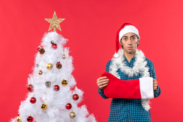圣诞老人一个戴着圣诞老人帽子 穿着蓝色条纹衬衫 手拿圣诞袜子的悲伤的年轻人 站在红色圣诞树旁帽子年轻人袜子