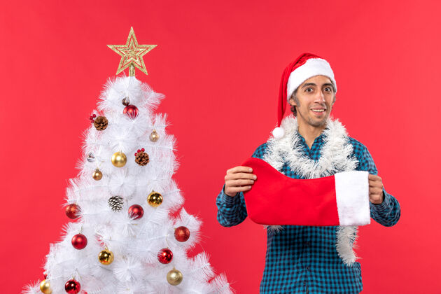 衬衫一个精力充沛的微笑着的年轻人 戴着圣诞老人的帽子 穿着一件蓝色的衬衫 手里拿着圣诞袜子袜子圣诞老人庆祝