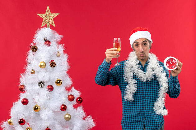 葡萄酒决心情绪化的年轻人戴着圣诞老人的帽子 拿着一杯葡萄酒和一个红色圣诞树旁的钟玻璃礼物时钟