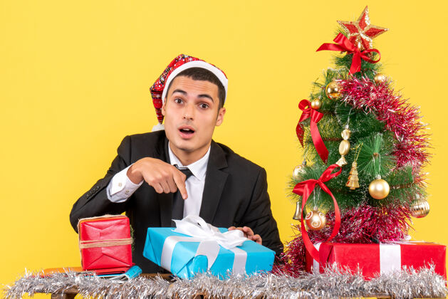 圣诞节前视图惊讶的男子与圣诞帽坐在桌上显示他目前的圣诞树和礼物坐职业展示