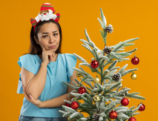 怀疑身穿蓝色上衣的年轻女子头戴滑稽的圣诞环 站在橙色背景下的圣诞树旁 带着怀疑的表情看着摄像机年轻圣诞搞笑