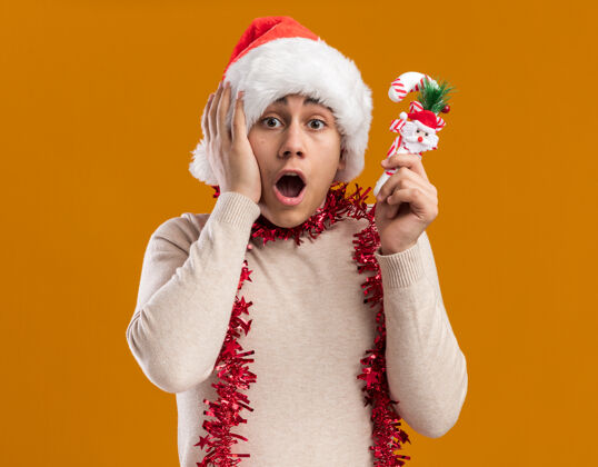 年轻被吓坏了的年轻人戴着圣诞帽 脖子上戴着花环 手里拿着圣诞糖果 手放在头上 被隔离在黄色的墙上头糖果圣诞节