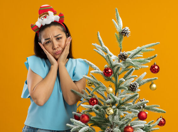 年轻身穿蓝色上衣 头戴滑稽圣诞环的年轻女子站在橙色背景下的圣诞树旁 神情悲伤地看着摄像机表情心烦圣诞