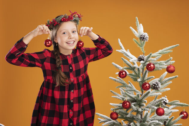 球快乐的小女孩戴着圣诞花圈 穿着格子衬衫 手里拿着圣诞球有橙色乐趣
