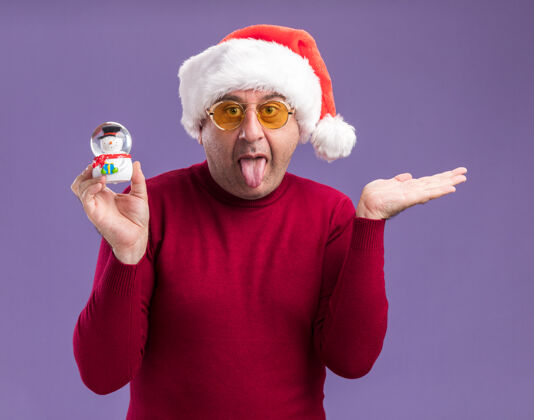 眼镜滑稽的中年男子戴着圣诞帽戴着黄色眼镜拿着圣诞雪球伸出舌头举起胳膊站在紫色背景上圣诞节中间舌头