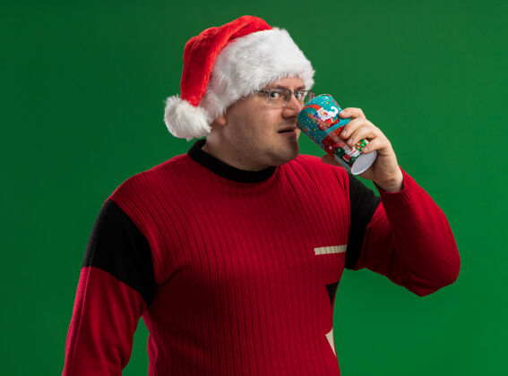 快乐令人印象深刻的成年男子戴着眼镜和圣诞老人的帽子喝咖啡从圣诞咖啡杯看着隔离在绿色背景上的相机眼镜印象深刻圣诞快乐