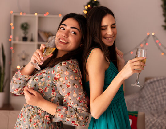 女孩笑容可掬的年轻姑娘们背靠背地站着 手里拿着香槟酒杯 在家里享受圣诞节时光年轻回来香槟