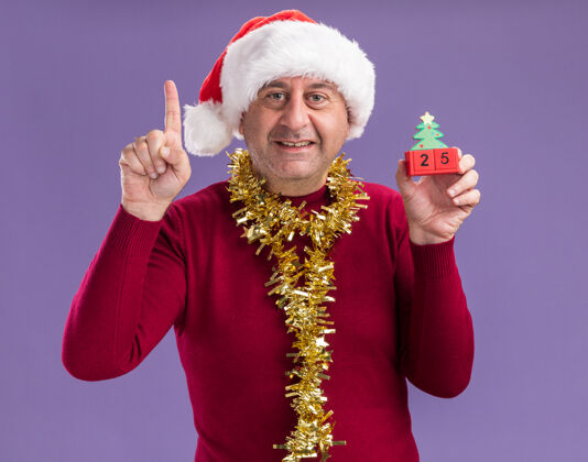 日期中年男子戴着圣诞老人帽子 脖子上戴着金属丝 手里拿着玩具方块 日期是25岁 看着相机微笑着 食指站在紫色背景上金属片五站着