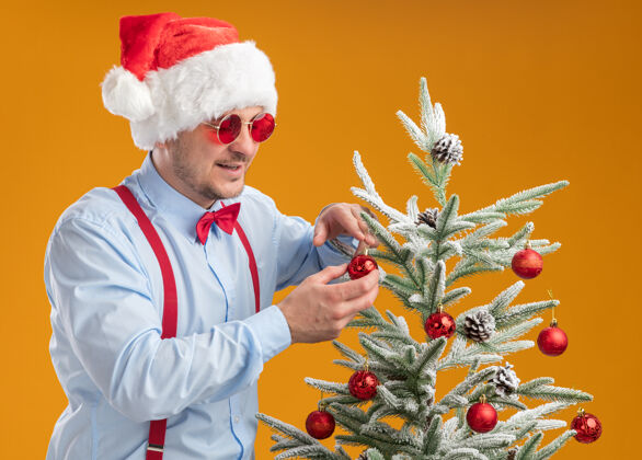 近一个戴着圣诞帽 戴着红眼镜 系着吊带领结的年轻人站在圣诞树旁 快乐地把玩具挂在树上 满心欢喜地挂在橘色的墙上站立玩具帽子