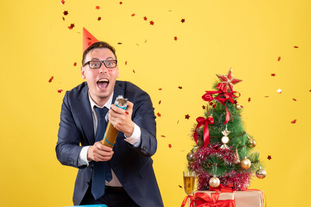 快乐前视图的快乐商人使用党波普尔站在桌子后面靠近圣诞树和黄色礼物表演者生意新郎