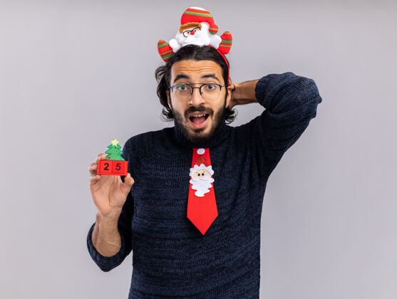 头发惊讶的年轻帅哥戴着圣诞领带 戴着发箍 手里拿着圣诞玩具 手放在脑后 隔离在白色背景上头惊讶手