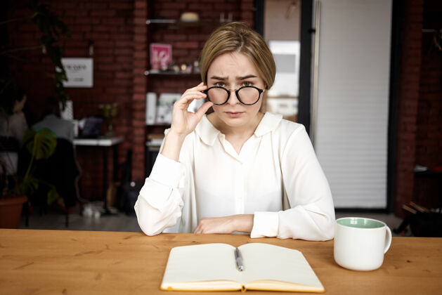 坐着严肃漂亮的年轻女雇主 穿着白衬衫 戴着眼镜 坐在办公室里 打开日记 在木桌上喝着咖啡 进行工作面试生意和事业经理工人工作场所