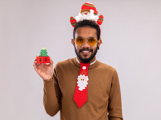 非洲快乐的非洲裔美国人 穿着棕色毛衣 头戴圣诞老人圈 打着有趣的红色领带 手里拿着玩具方块和25岁的约会对象 站在白色背景下微笑着看着相机搞笑立方体举行
