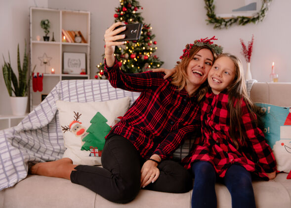 看带着冬青花环的笑妈妈和女儿看着手机坐在沙发上自拍 在家里享受圣诞节时光女儿电话自拍