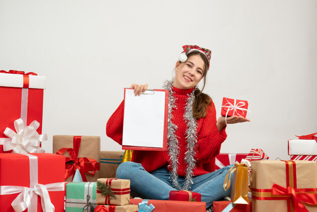 盒子戴着圣诞帽的快乐女孩拿着礼物和文件坐在白色的礼物旁边购物帽子快乐