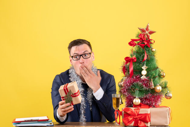桌子前视图惊讶的人拿着礼物坐在圣诞树旁边的桌子上 黄色的礼物人表演者圣诞节