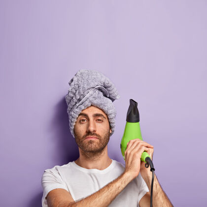 干燥自信的没刮胡子的男人竖起电扇 头上戴着毛巾 去干头发垂直护理吹风机