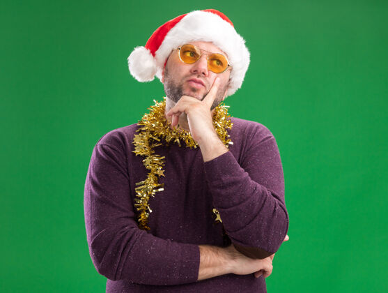侧面体贴的中年男子戴着圣诞帽 脖子上戴着金箔花环 戴着眼镜 手放在下巴上 看着隔离在绿色背景上的一边 还有复印空间圣诞老人周围脖子