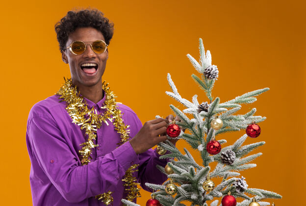 金属片顽皮的美国黑人年轻人戴着一副戴着金箔花环的眼镜 站在圣诞树旁 用圣诞饰品装饰圣诞树新年脖子眼镜