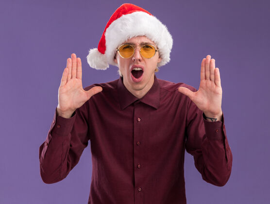 保持一个戴着圣诞帽戴着眼镜的金发年轻人看着摄像机双手举在空中大声呼喊着紫色背景上孤立的东西大声金发喊叫
