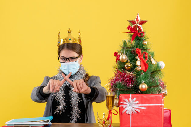 十字架前视图：戴着医用面罩的年轻女孩交叉手指 圣诞树和鸡尾酒礼物面具桌子医学