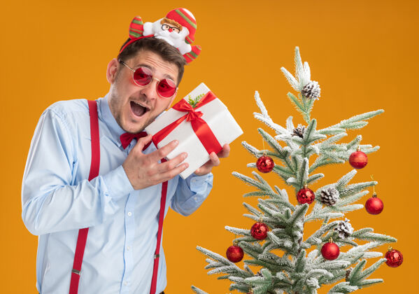 领带戴着吊带领结的年轻人站在圣诞树旁 戴着圣诞老人和红眼镜 手里拿着一份礼物 在橙色的背景下快乐而兴奋举行年轻圣诞老人