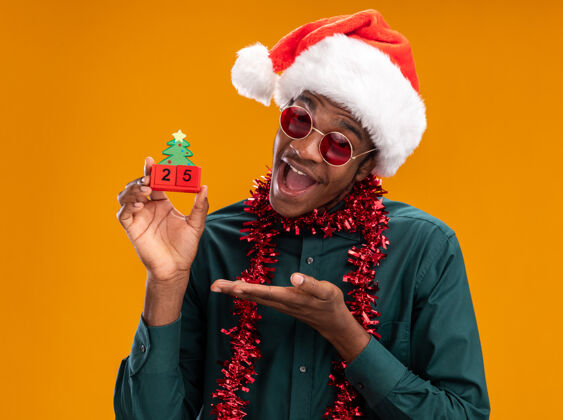 提出快乐的非洲裔美国人戴着圣诞帽 戴着花环 戴着太阳镜 手里拿着日期为25的玩具立方体 手臂站在橙色背景上五非洲举行