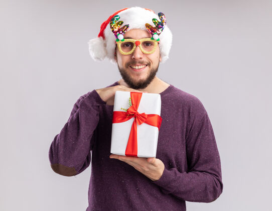 圣诞老人快乐快乐的年轻人穿着紫色毛衣 戴着圣诞帽 戴着滑稽的眼镜 拿着礼物 站在白色的背景下 面带微笑地看着镜头毛衣帽子抱着