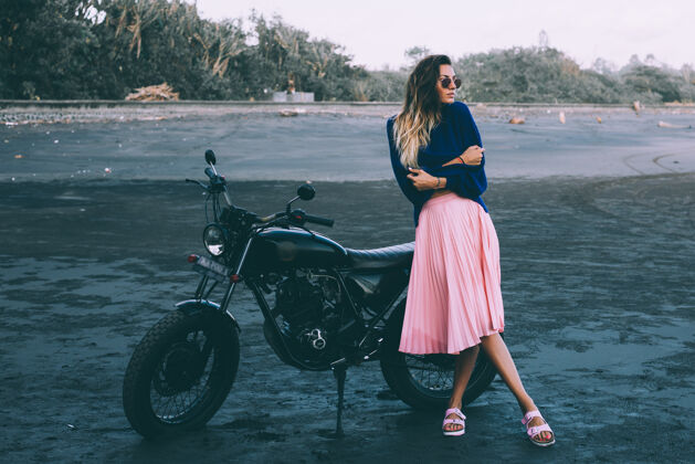 车辆戴着太阳镜 穿着蓝色毛衣和粉色裙子的时髦女人的时尚肖像 站在黑色沙滩上的黑色自行车旁女人自行车骑自行车的女孩