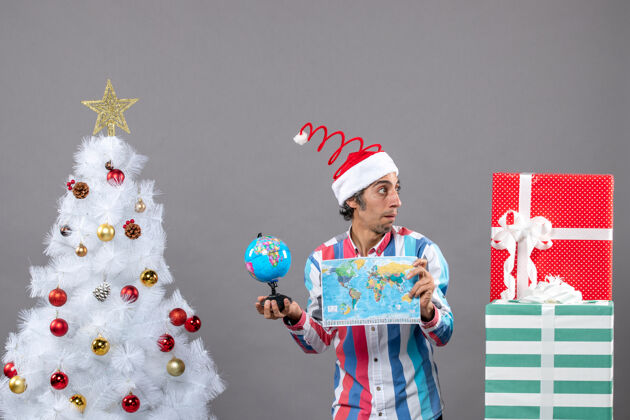 礼物正面图惊讶的男子戴着螺旋弹簧圣诞帽看着礼物拿着世界地图和地球仪春天快乐男人