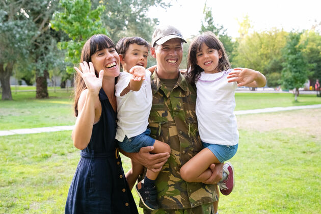 孩子一对幸福的家庭夫妇和两个孩子在公园里合影军人抱着孩子 他的妻子拥抱他们并挥手中镜头家庭团聚或回家的概念男人摆姿势团结